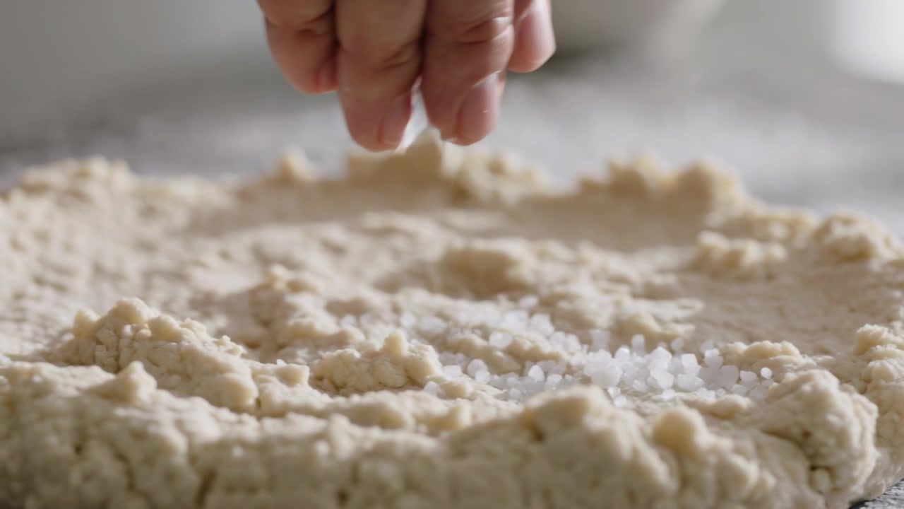Sprinkling cook's bare hand pinching coarse salt across flat dough on floured countertop, seasoning