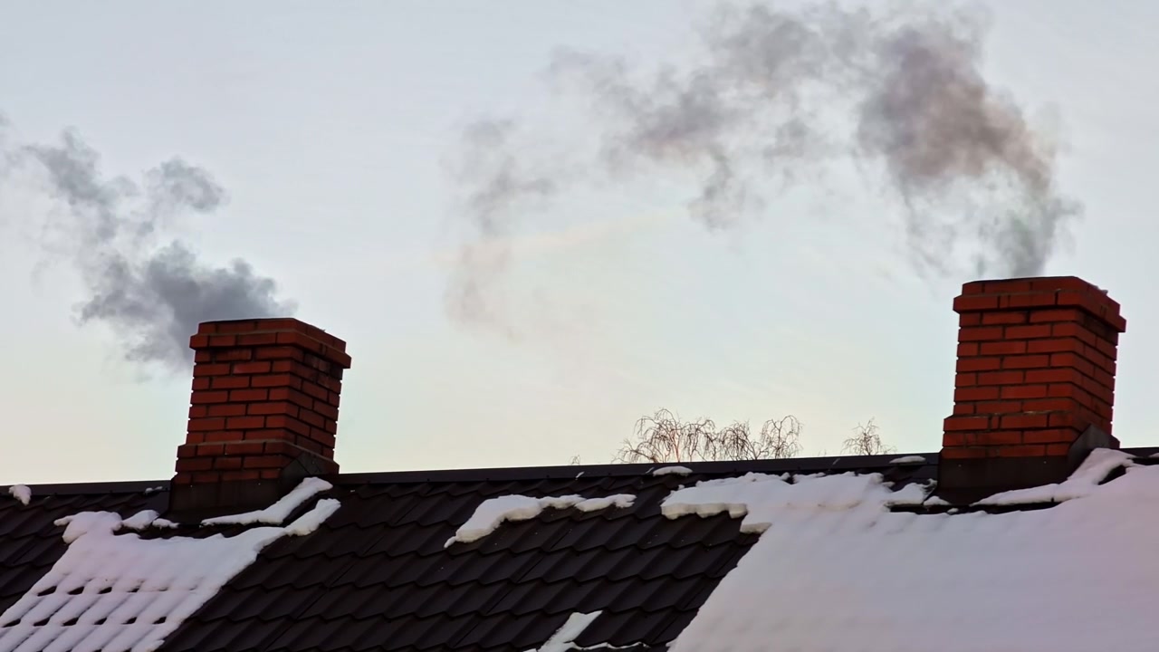 Dark smoke billowing from two brick chimneys on a house roof covered with snow in winter