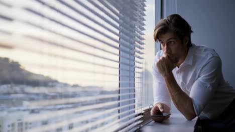 Front-view-of-young-Caucasian-male-executive-with-hand-on-chin-leaning-at-window-in-modern-office-4k