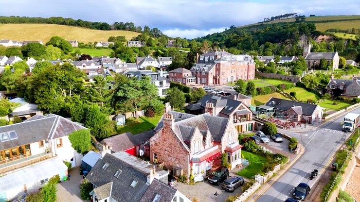 Drone footage glides above Rosemarkie, Highland, Scotland, revealing residential houses, historic buildings, and lush greenery under bright daylight with smooth camera movement