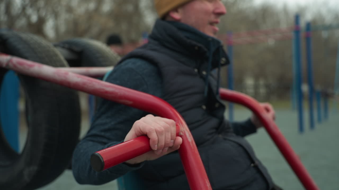 A coach wearing a brown beanie and black jacket is working out on outdoor equipment, performing pull-ups with red iron bars, after completing the pull-ups, he starts rotating his arms