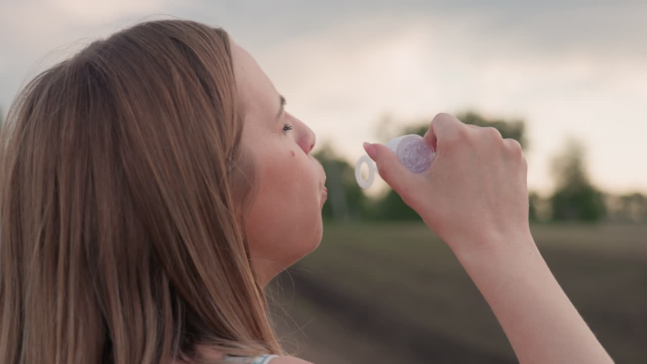 close up side view of woman blowing soap bubbles with handheld wand in open countryside field during sunset, delicate iridescent spheres drifting gently against soft pastel sky