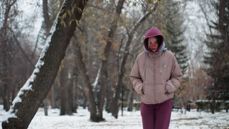 lady jumps playfully in winter snow park, dressed in warm hoodie and coat, snow-covered trees in background create a peaceful, scenic atmosphere