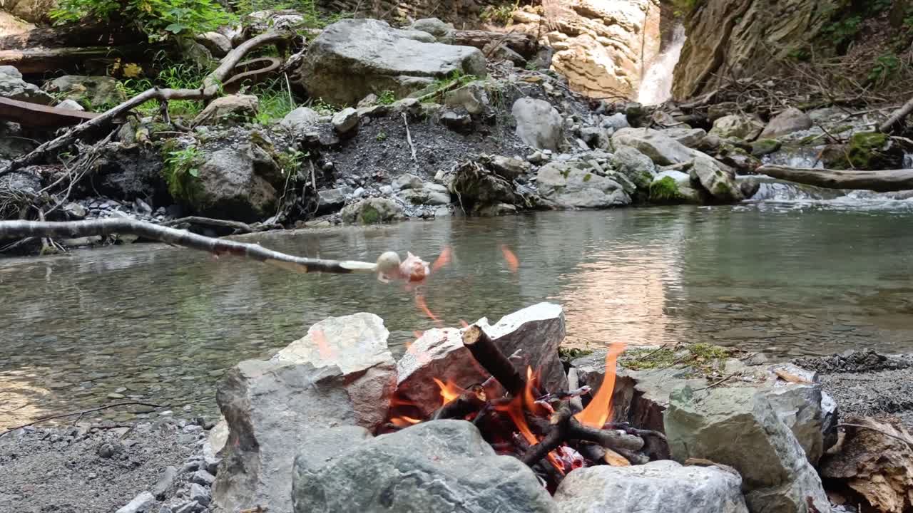 Uomo che cucina salsicce su un bastone sul falò con cascata e fiume sullo sfondo