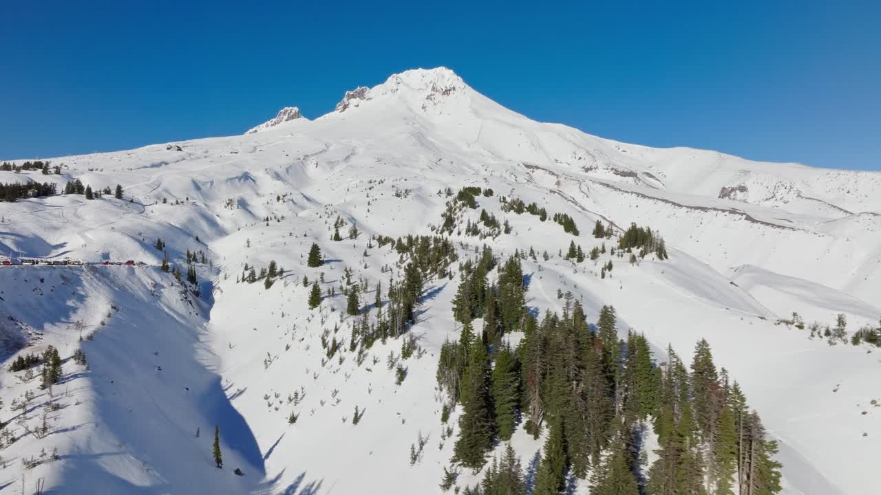 Bidikan drone udara panning dari Gunung Hood yang tertutup salju dan gletsernya, fumarol, dan jurang di dekat Timberline Resort yang terkenal di dekat Portland Oregon saat matahari rendah terbenam di musim dingin dengan pohon pinus