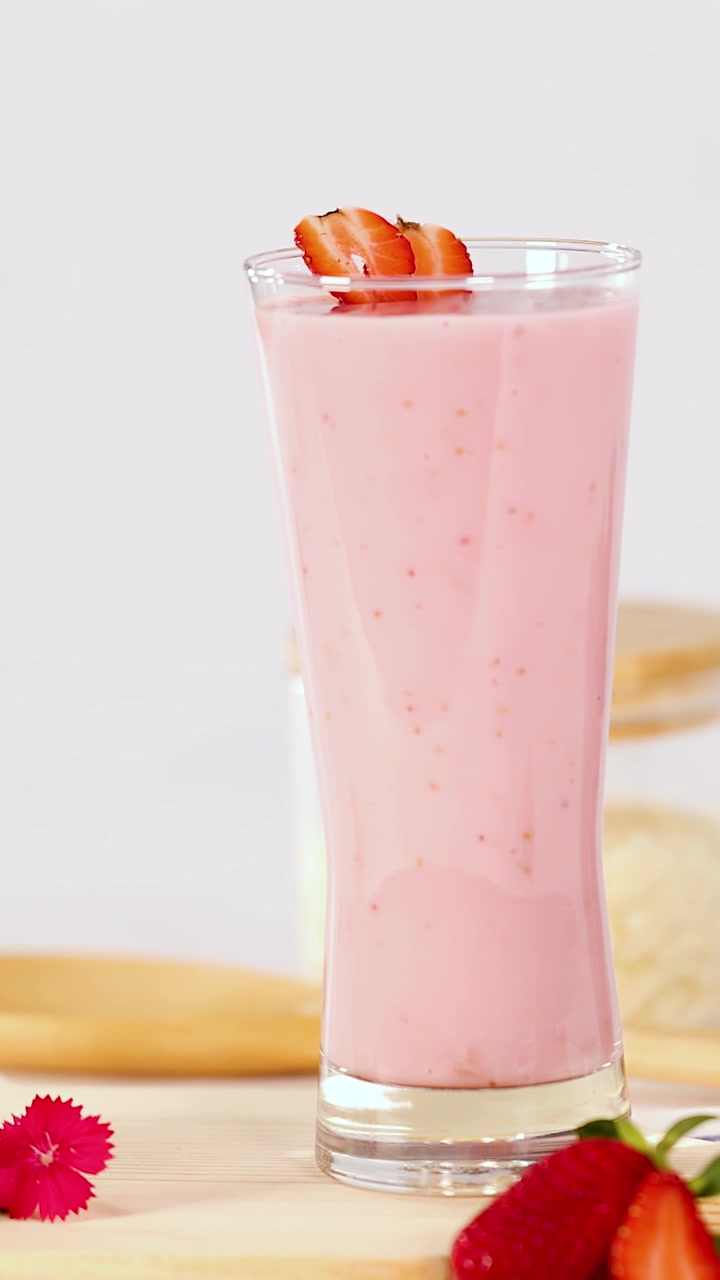 Una mano adagia fragole a fette su un cremoso frullato rosa in un bicchiere alto, sotto una luminosa luce naturale.