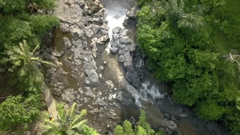 Luftneigung-Nach-Unten-über-Den-Wasserfall-Von-Bali,-Erstaunlicher-Flusslauf-Zum-Tegenungan-Wasserfall,-Indonesien,-Mit-Umliegenden-Felsen