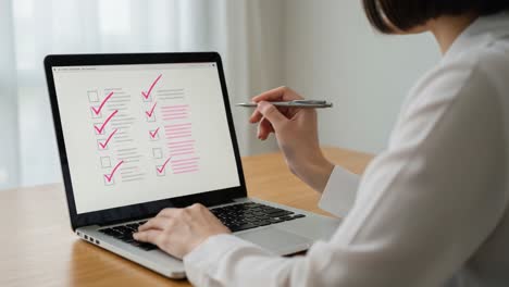 a focused individual engaging in detailed work on a laptop, methodically reviewing a checklist with visible marks of completion in a bright office setting