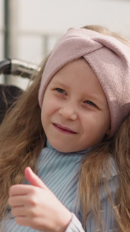 little girl with muscular weakness turns to camera trying to show thumb up gestures. curly-haired child sits in wheelchair on blurred background closeup