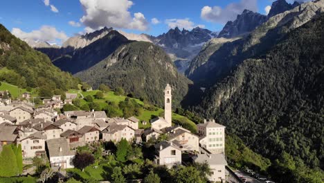 Sobrevuelo-Aéreo-En-El-Sol-De-La-Tarde-Sobre-El-Histórico-Pueblo-De-Soglio-En-La-Región-De-Bregaglia-De-Grissons,-Suiza-Con-Vistas-A-La-Antigua-Iglesia-Y-A-Los-Picos-Montañosos-De-Engadin