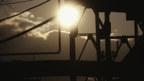 Silhouette-of-an-industrial-structure-against-a-backdrop-of-a-bright-sun-and-clouds,-creating-a-dramatic-and-contrasting-visual-effect