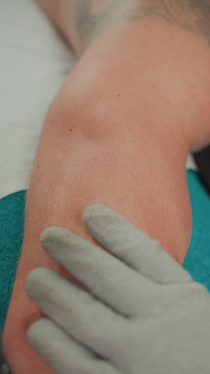 Massaggio a microcorrente con guanto sull'avambraccio tatuato, terapista che leviga la pelle con guanto conduttivo testurizzato, passaggi delicati intorno al gomito, sottili particelle esfolianti, ambiente spa clinico con asciugamano color ottanio.