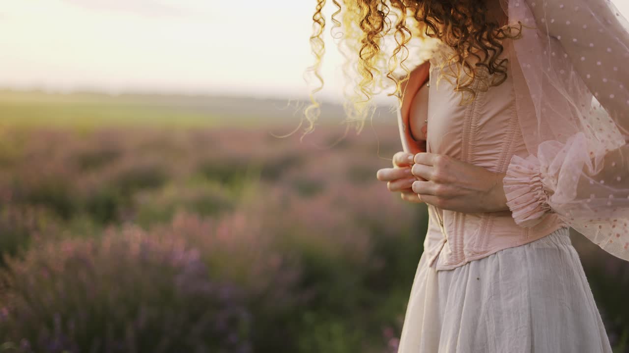 Ragazza dai capelli ricci in un vestito rosa, stringe il suo corsetto in mezzo a un campo di lavanda viola.