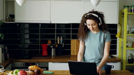 young teenage girl wearing headphones appears in kitchen coming to table, drinking tea from cup, dancing and printing something on laptop