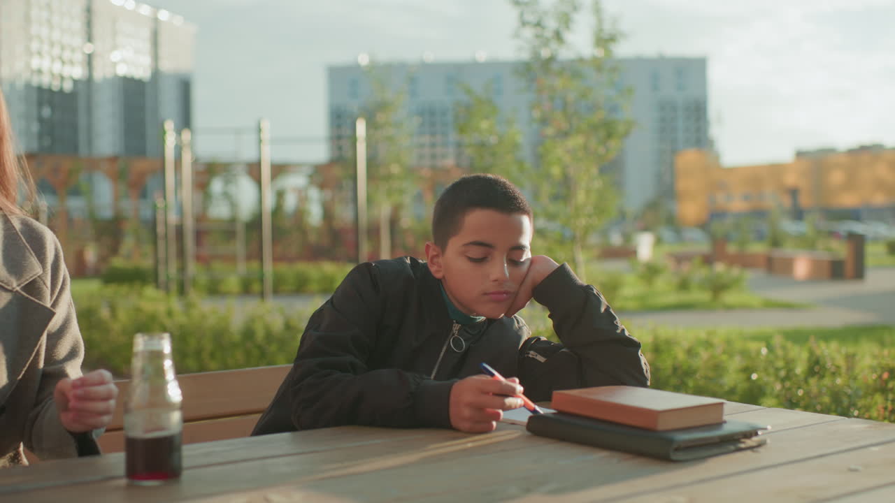 Boy writing calmly on wooden table outdoors with book and pen while woman takes chips from bag to offer him, highlighting everyday care, support, and family connection in urban park setting