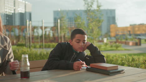 boy writing calmly on wooden table outdoors with book and pen while woman takes chips from bag to offer him, highlighting everyday care, support, and family connection in urban park setting