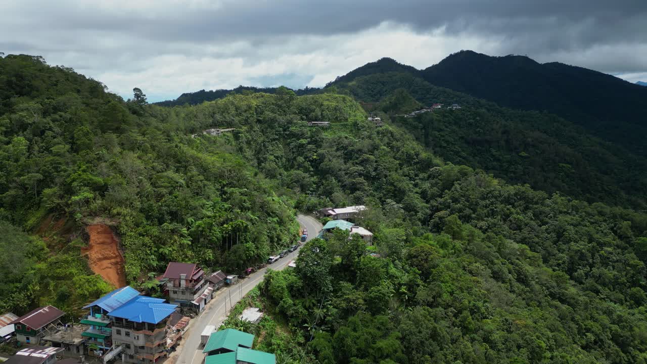 Gliding aerial shot above the main road and clustered houses in the mountainous, heavily forested terrain of Banaue, Ifugao, Philippines, under a cloudy sky