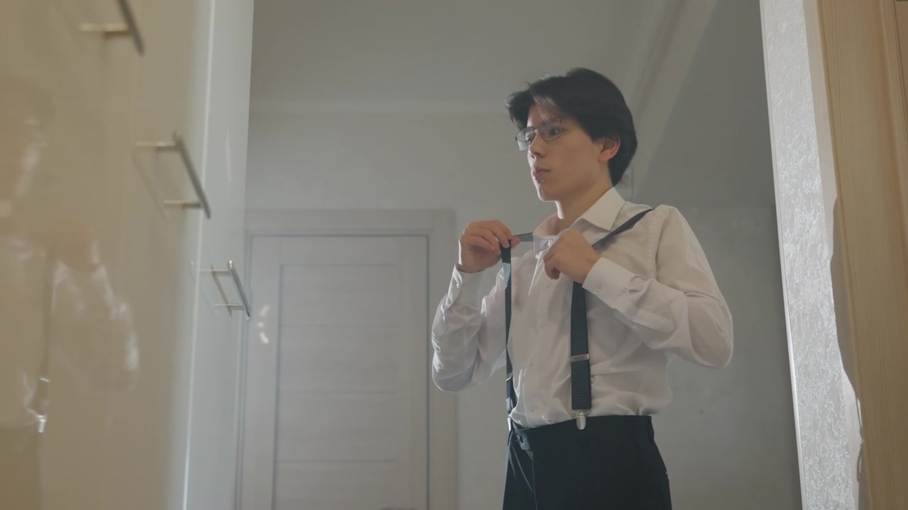 Uomo asiatico che si sistema le bretelle nel corridoio. Luce naturale soffusa attraverso la porta, camicia bianca e pantaloni sartoriali, rituale silenzioso prima di un evento importante, umore introspettivo con inquadratura cinematografica, opzioni.