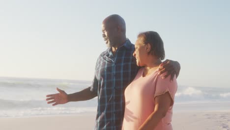 Smiling-senior-african-american-couple-embracing-and-walking-on-sunny-beach