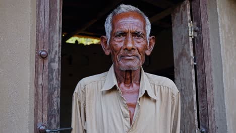 elderly man with gray hair and weathered skin stands in the doorway of a simple dwelling, his gaze meeting the viewer's, conveying a sense of quiet dignity and experience