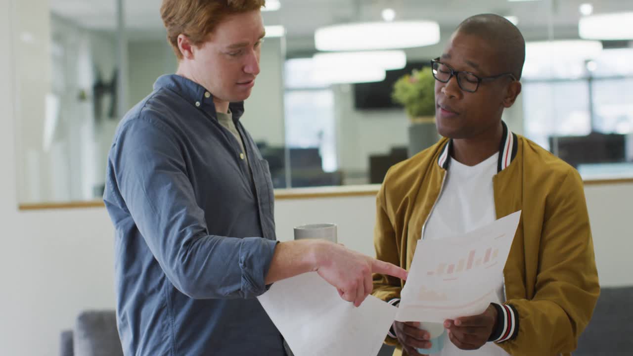 Two diverse male colleagues looking at charts, having business talk in office