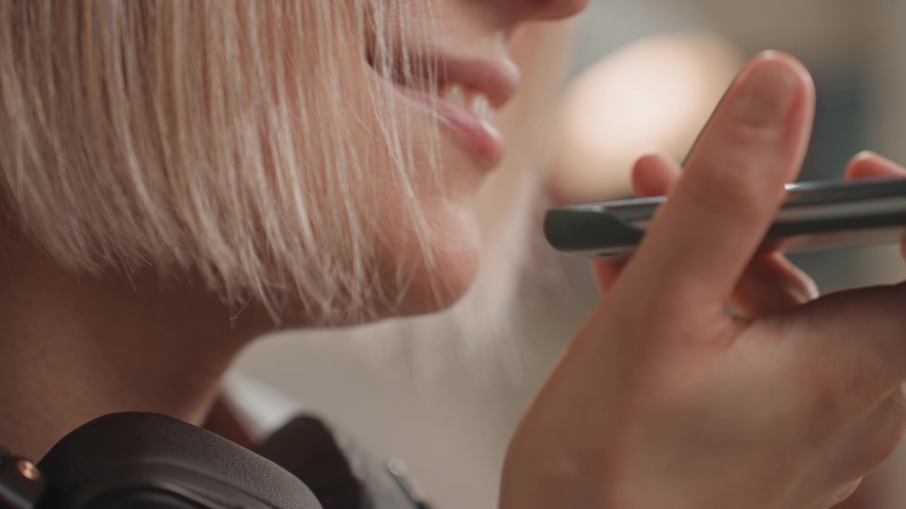 Primo piano di una donna bianca che parla al telefono, memo vocale dolce e intimo per l'amante, cuffie intorno al collo, luce calda interna, sorriso gentile, sussurro affannoso, registrazione per una demo musicale, atmosfera spontanea da studio casalingo