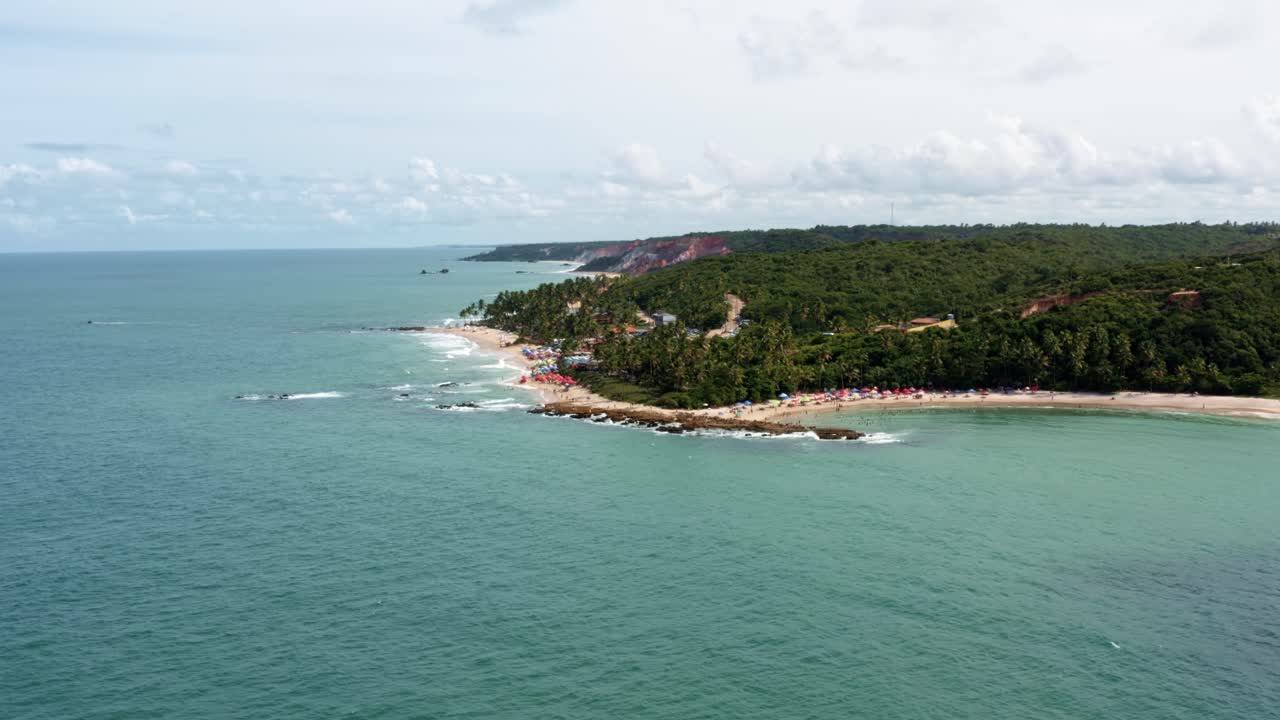 dolly en un dron aéreo toma amplia de la popular playa tropical de coquerinhos cubierta de paraguas con turistas nadando en una piscina natural de un arrecife en condé, paraíba, brasil