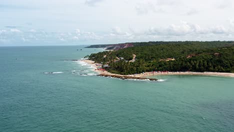 dolly in aerial drone wide shot of the popular tropical coquerinhos beach covered in umbrellas with tourists swimming in a natural pool from a reef in conde, paraiba, brazil