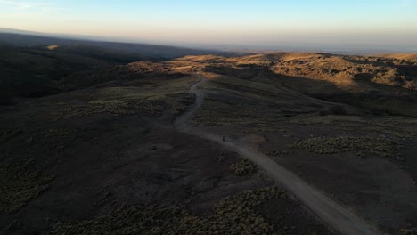 Toma-Aérea-De-Una-Motocicleta-Conduciendo-Por-Un-Camino-De-Montaña-Accidentado-Durante-La-Puesta-De-Sol