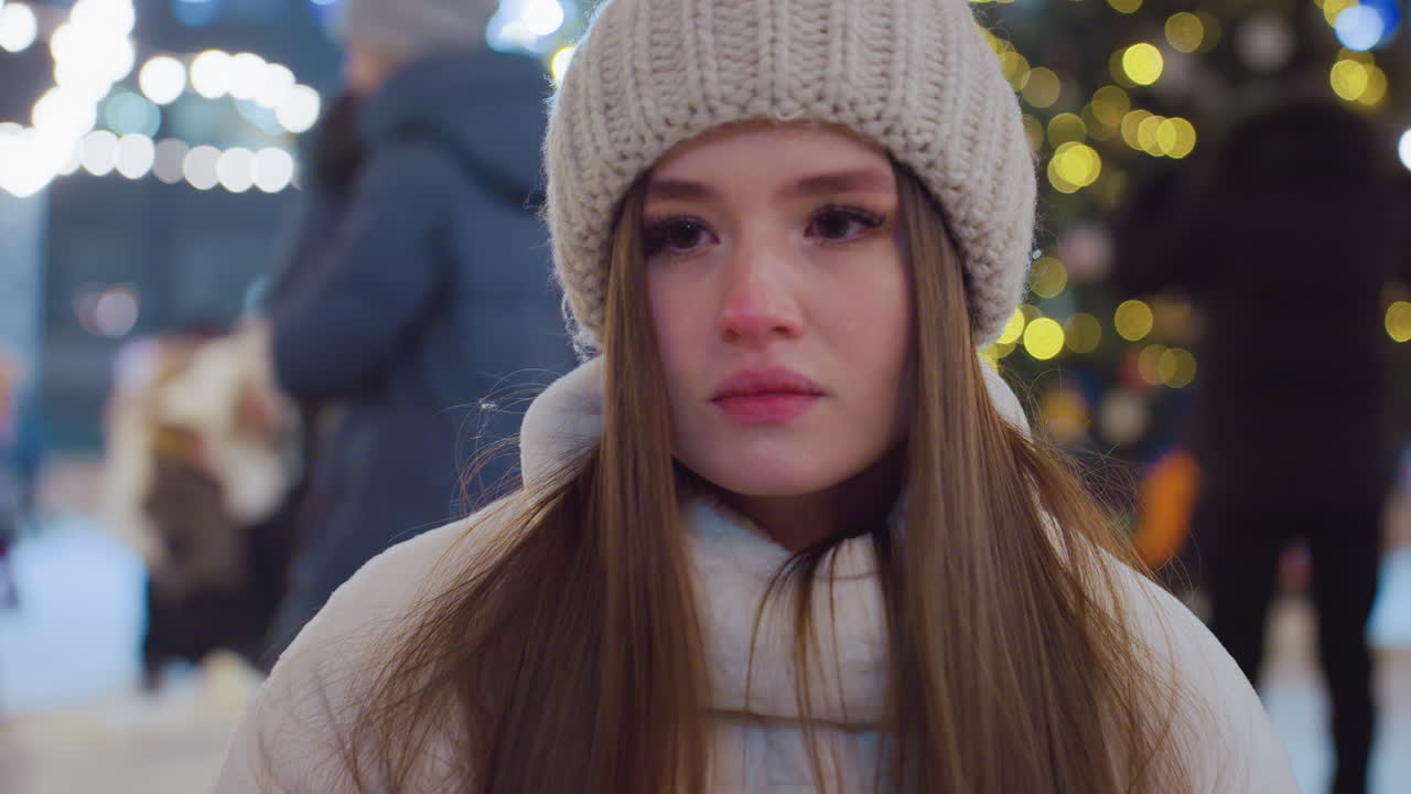 Young woman in warm winter outfit sitting outdoors at night, lost in thought, festive lights illuminate the scene as people move around, skating and enjoying the lively holiday atmosphere in the city