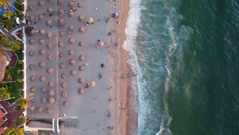 Touristen-Am-Strand-Von-Los-Muertos-Mit-Strohschirmen-Bei-Sonnenuntergang-In-Puerto-Vallarta-Mexiko,-Antenne-Von-Oben-Nach-Unten