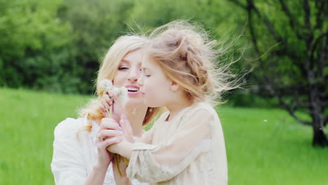 cool blonde girl blows a dandelion with her mother in a green forest