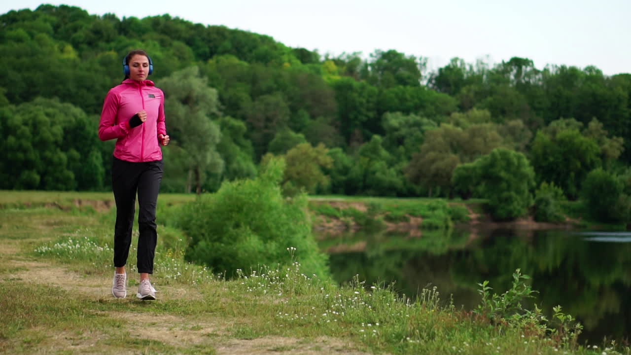 A girl in a pink jacket and black pants runs near the river in headphones preparing for the marathon