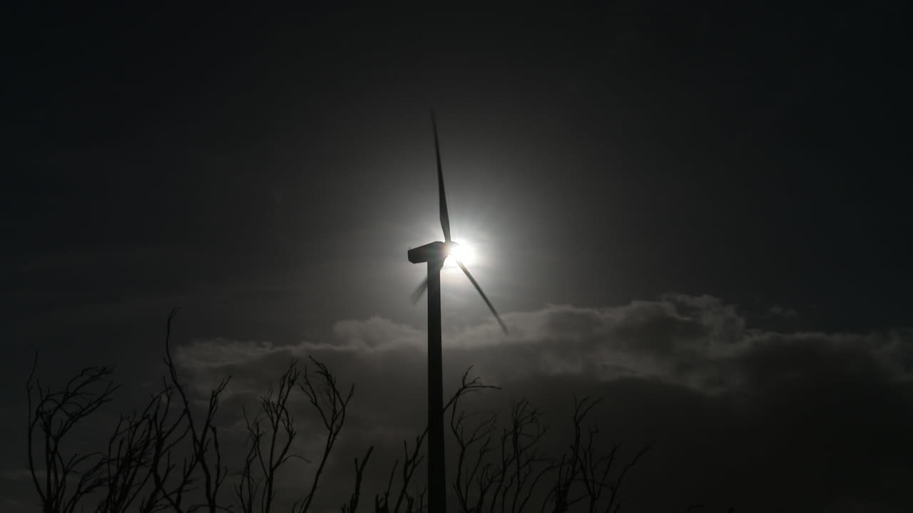 "A stunning night drone shot of a wind turbine gracefully rotating under the stars. Its blades, subtly illuminated, cut through the darkness as the structure silently generates clean, renewable energy