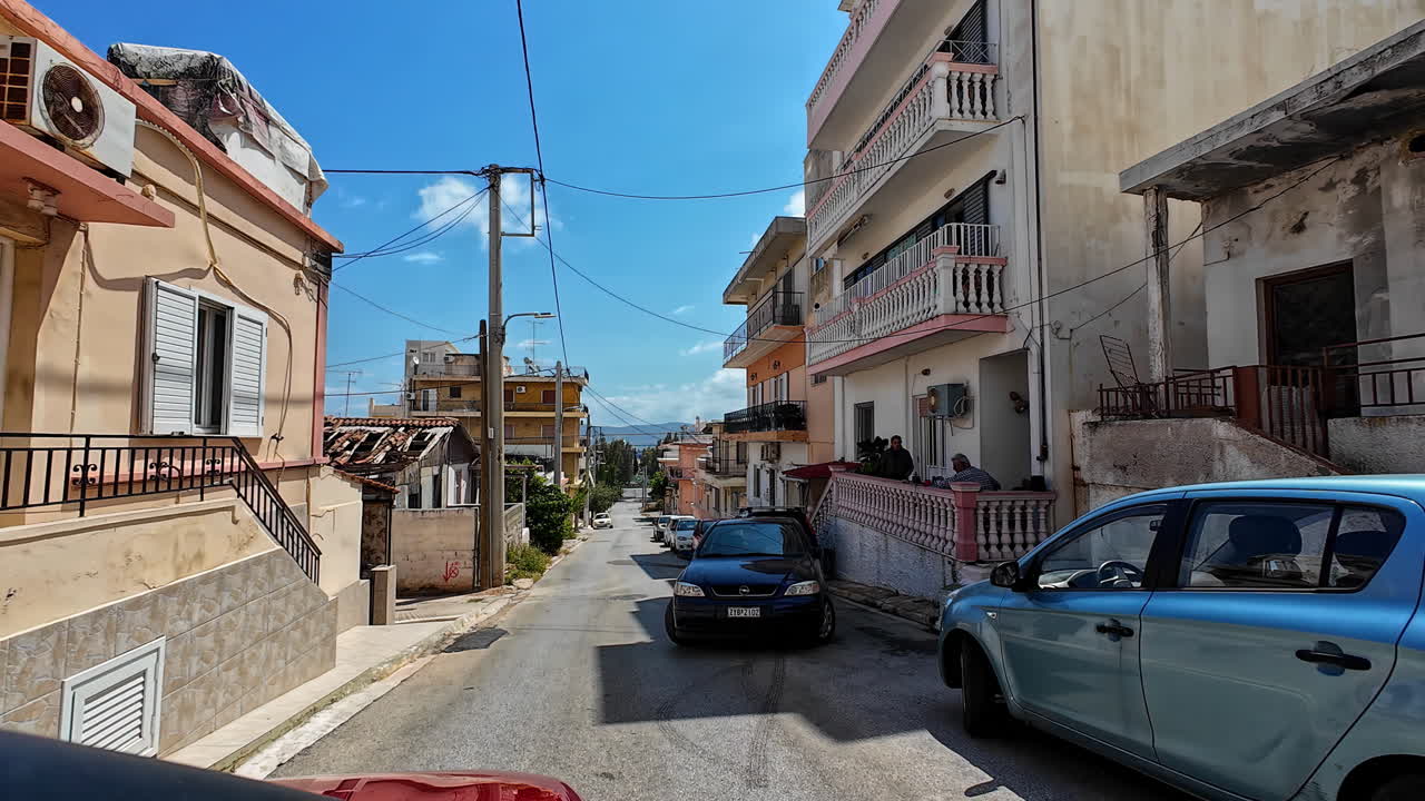 A sun-drenched street in a Greek town with residential buildings and parked cars.