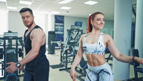 Smiling couple doing workout in fitness club. Beautiful female trainer shows different exercises to a man in front of the mirror at gym indoors.