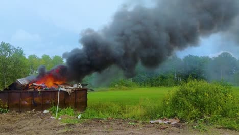 thick dark smoke billowing from a burning dumpster full of garbage on rural farmland