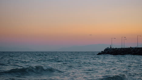 Evening-waterscape-with-wavy-sea-rocky-pier-and-flying-gull