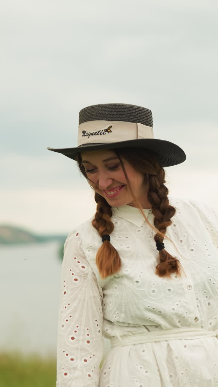 한 여자가 뒷면에 보이는 호수와 함께 초록 잔디에 서 있습니다. 그녀는 세련된 모자와 복잡한 패턴의  드레스를 입고, 으로 보면서 재미있는 얼굴을 하고 있습니다.