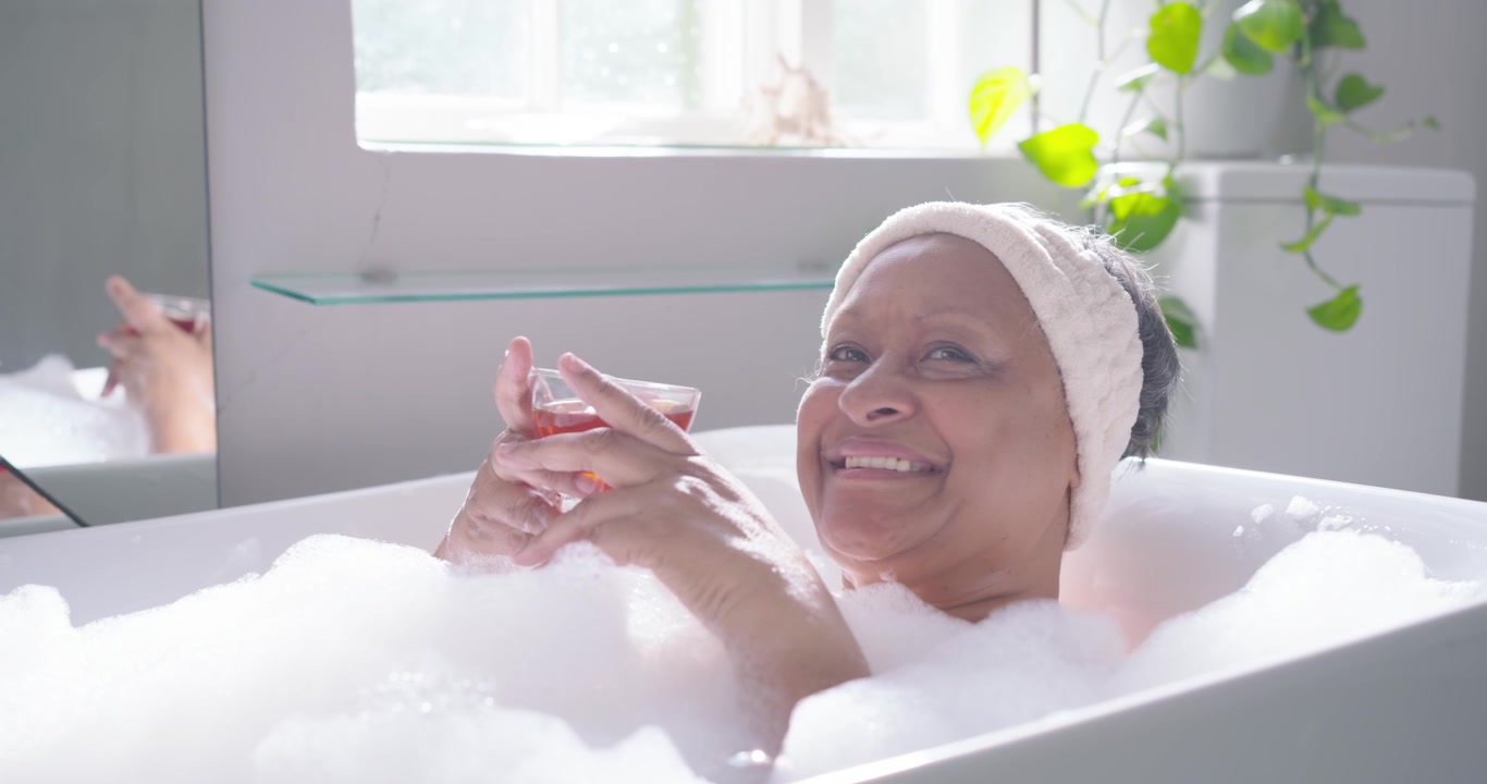 Senior African American woman reclining in modern tub with foam, lifting glass, sipping to relax