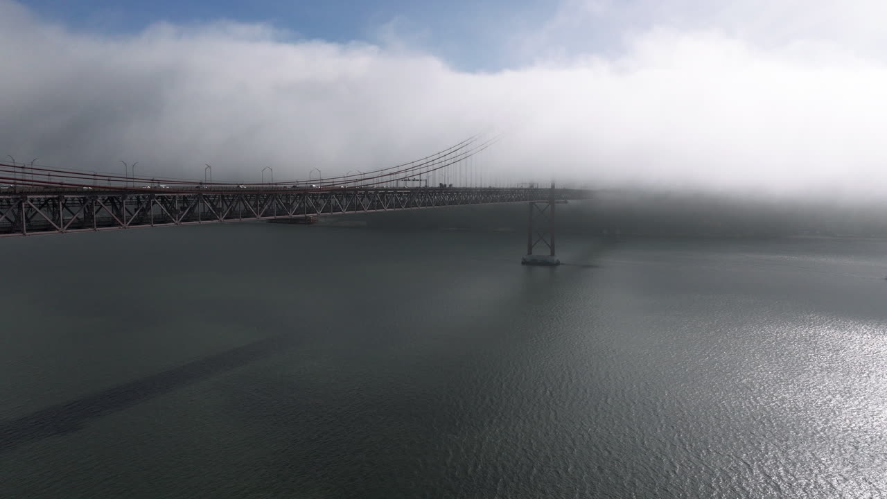 Aerial drone shot towards the 25th April Bridge in Lisbon, Portugal, Europe. Sunny and bright with clouds, fog, mist, covering the bridge
