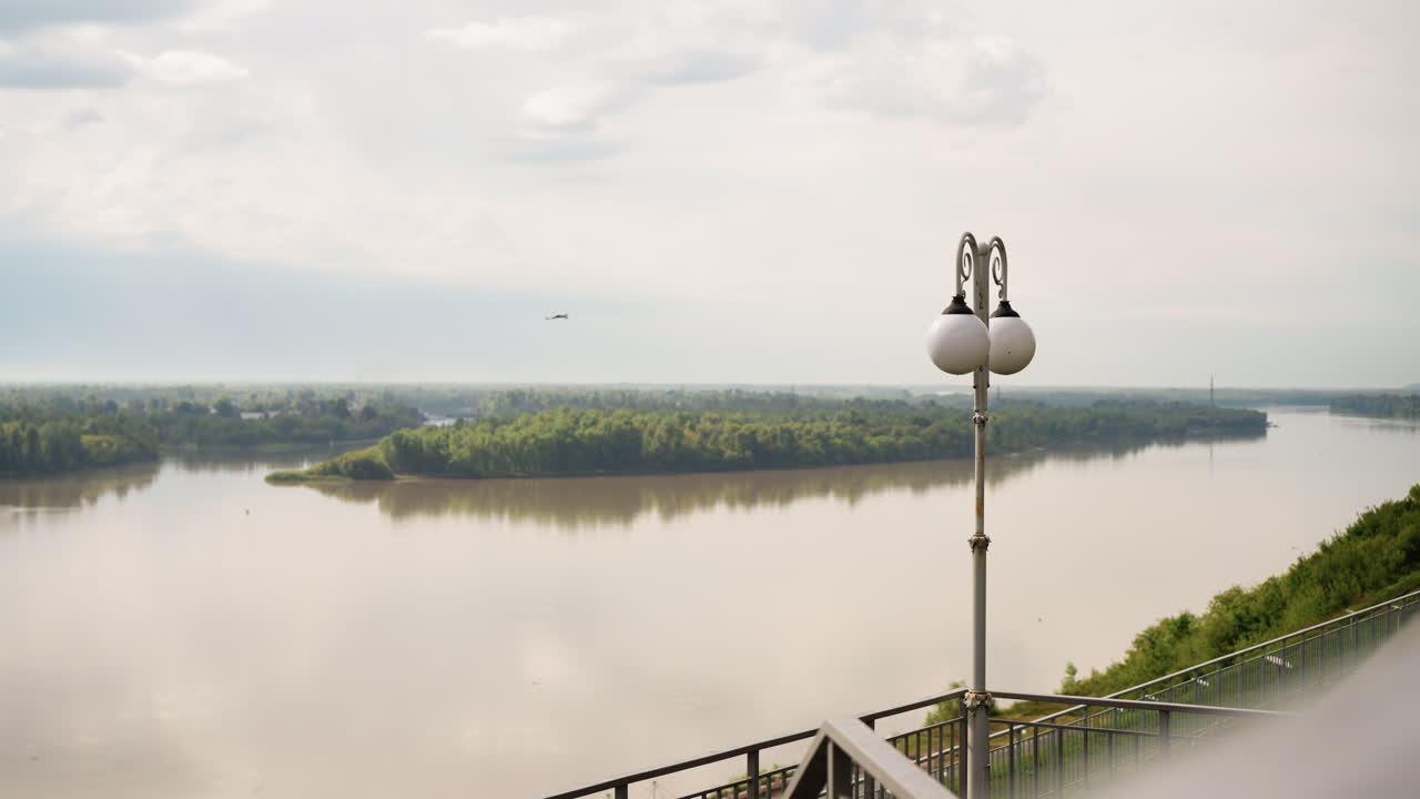 가로등과 섬이 있는 강가 전망, 부드러운 구름이 있는 하늘과 잔잔하게 반사되는 물, 멀리 나무가 덮인 둑, 전경에 보이는 철제 난간과 난간 동자, 은은한 안개와 흐린 분위기, 섬세함