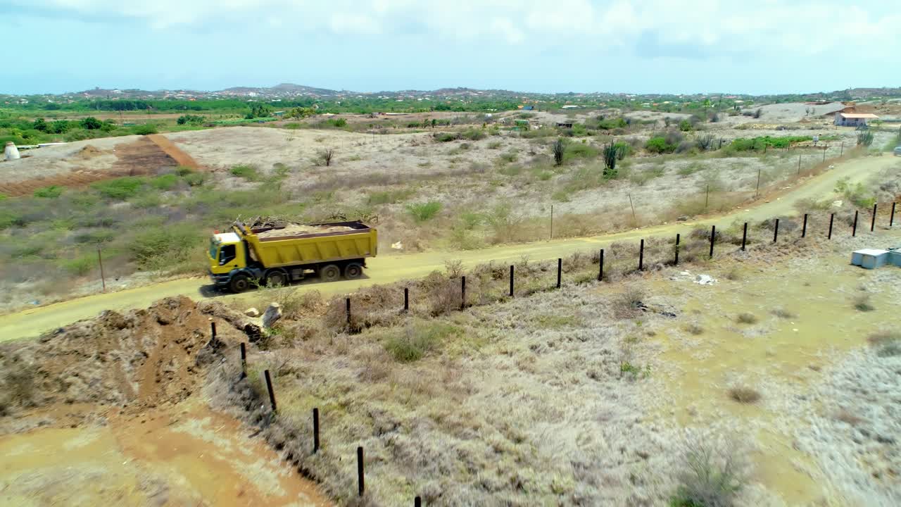 4k de inclinación cinematográfica aérea hacia abajo de un camión de basura amarillo lleno de tierra y basura conduciendo a través de tierras de cultivo rurales en un camino de tierra durante un hermoso día soleado en el caribe, curacao