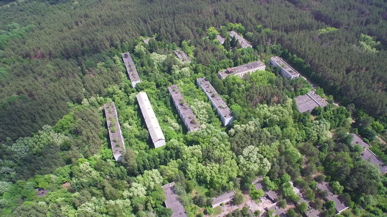 Drone footage of nature reclaiming abandoned buildings in Chernobyl, Ukraine. Trees grow on roofs, obscuring facades., Chernobyl-2 Military base