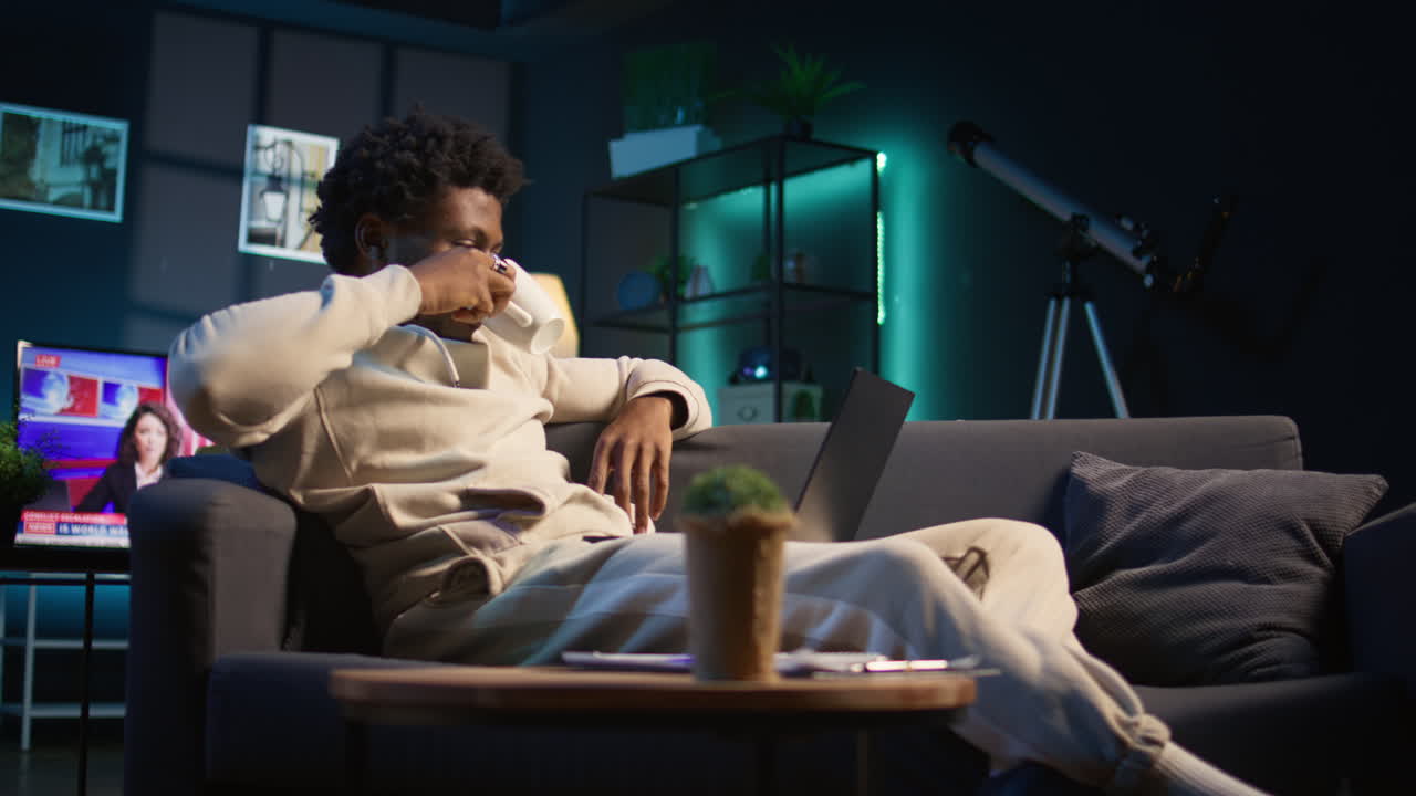 Casual person opening his laptop at home to solve daily tasks on his couch, doing remote freelance work online. Young black man surfing the web multitasking in loft apartment, tech savvy. Camera B.