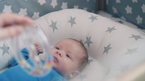 little child looks at rattle in mommy hand in cozy baby bed