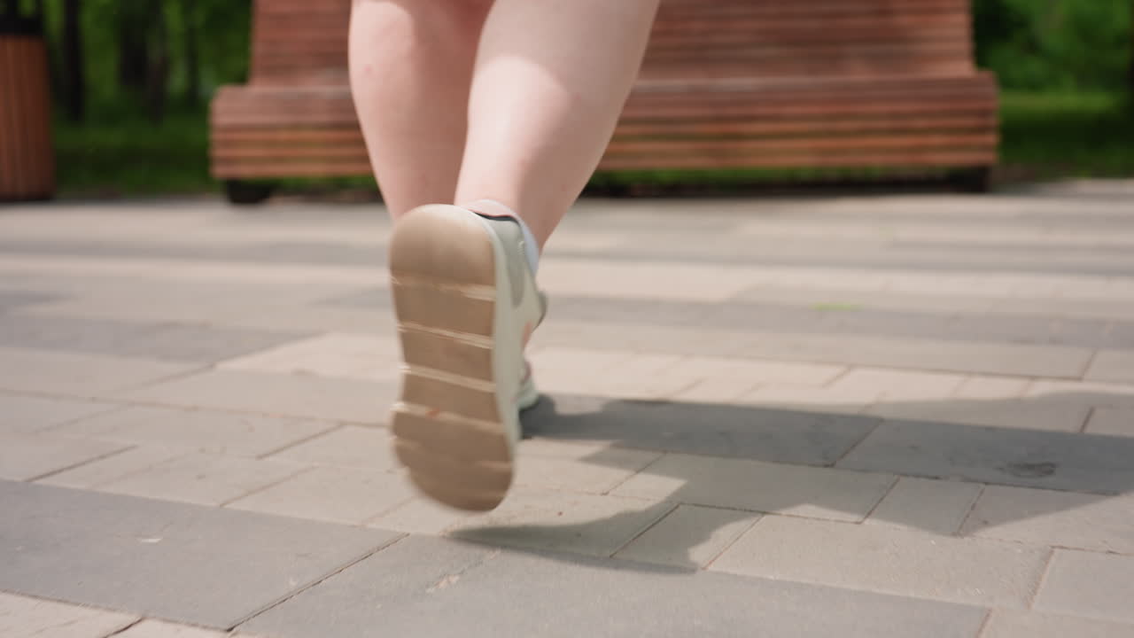 Close up view of woman legs in white sneakers walking on stone pavement toward wooden bench in sunny green park, gentle summer light creating soft shadows