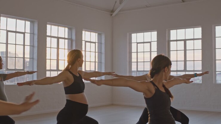 istruttore di lezione di yoga che insegna alle donne incinte sane che praticano la posa del guerriero godendo di esercizi di allenamento di gruppo in palestra all'alba