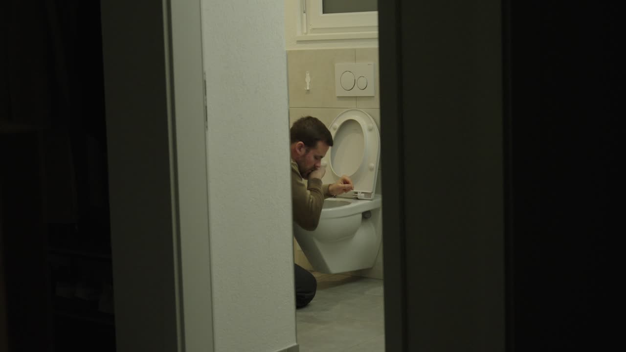 Nighttime scene of a man feeling nauseous while sitting on the toilet, viewed through a doorway. Concept of sickness, digestive issues, nighttime illness, and physical discomfort