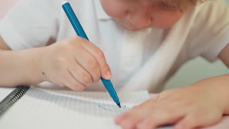 Little-boy-draws-on-notebook-page-with-blue-marker-at-table